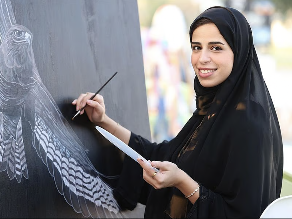 A smiling Emirati female artist wearing a black abaya and hijab paints a detailed black-and-white falcon on a canvas. She holds a paintbrush and palette, with a blurred outdoor event in the background.